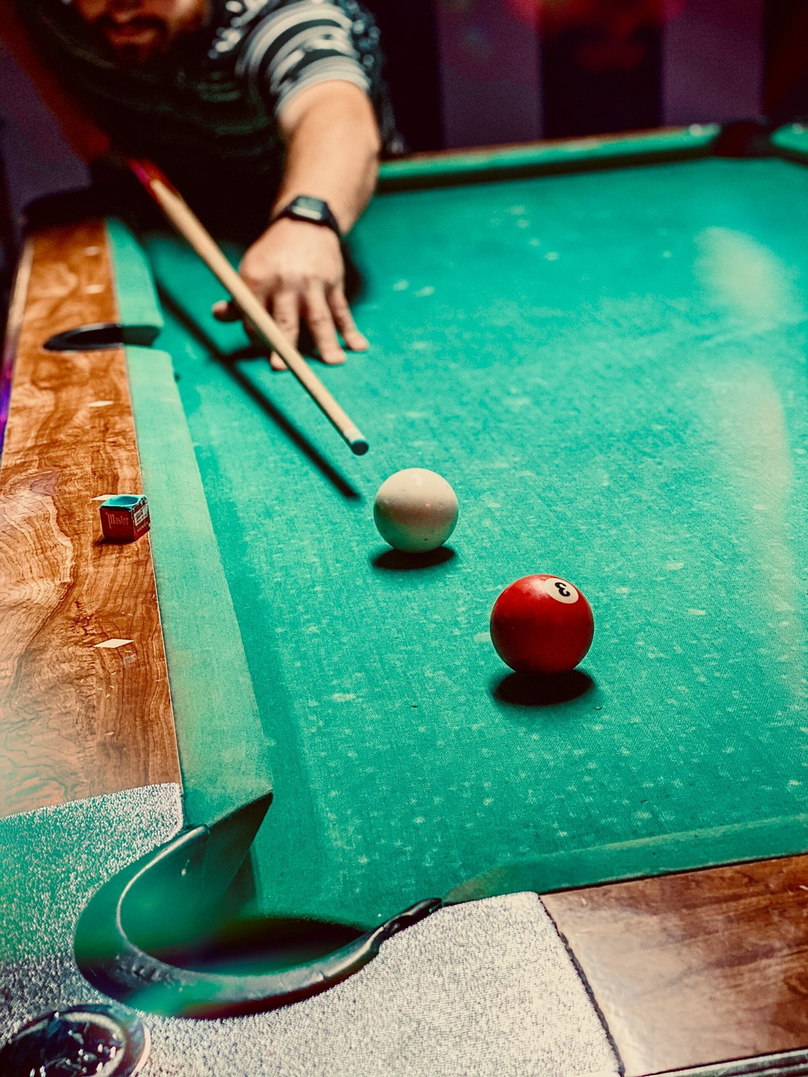 A person in a striped shirt aiming a cue stick to hit the cue ball on a green pool table, with the red 3-ball nearby and chalk on the table edge.